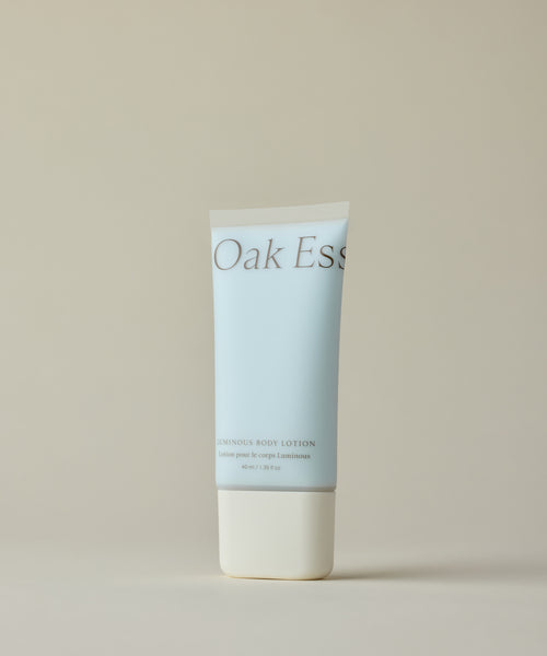 A light blue tube of Oak Ess Luminous Body Lotion Travel with Blue Tansy and a white cap stands upright on a beige background. The label shows minimalist text in soft brown and blue shades.