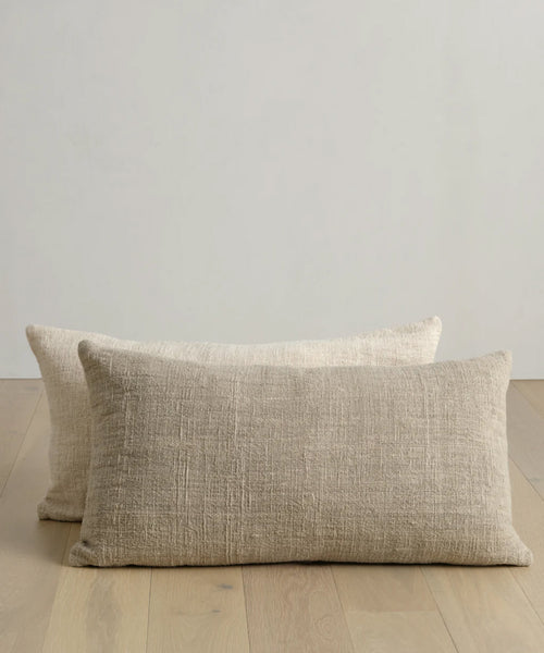 Two Luna Lumbar Pillows, handwoven in textured beige, are placed on a light wooden floor against a plain light wall, with one pillow slightly in front of the other.