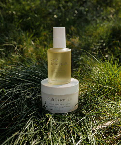 The Lymphatic Massage Set from Oak Essentials—featuring ritual body oil and perfecting body scrub—is displayed outdoors on green grass in natural sunlight, ideal for a rejuvenating massage ritual.