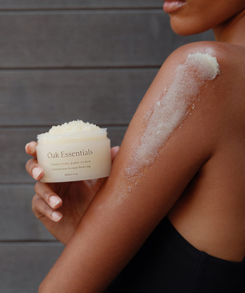 A person applies the Lymphatic Massage Set to their shoulder, holding the jar in one hand. The scrub is massaged onto their upper arm as part of a soothing massage ritual, set against a wooden background.
