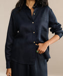 A person in a loose-fitting, dark navy Madeline Shirt with a chest pocket and matching pants stands with one hand in their pocket. The shirt’s linen texture highlights their long dark hair and bold green ring.