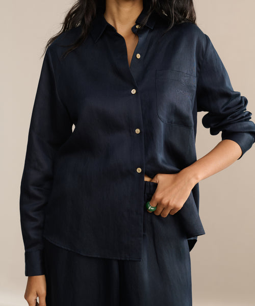 A person in a loose-fitting, dark navy Madeline Shirt with a chest pocket and matching pants stands with one hand in their pocket. The shirt’s linen texture highlights their long dark hair and bold green ring.