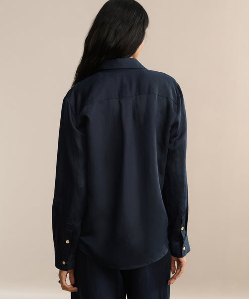 A person with long dark hair is seen from behind, wearing a loose-fitting, dark navy Madeline Shirt and matching pants with a subtle linen texture, set against a plain beige background.