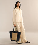 A woman wears the Madeline Shirt and matching wide-leg pants with a linen texture, holding a large black woven tote bag with a cane panel. She stands against a neutral background in black slip-on shoes.