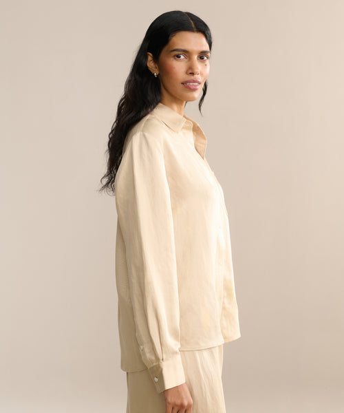 A woman with long dark hair stands sideways, looking at the camera against a plain beige background while wearing the Madeline Shirt and matching pants with a subtle linen texture, perfect for spring.