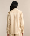 A person with long dark hair is seen from behind wearing the Madeline Shirt in light beige linen texture, paired with matching pants for a spring-ready look against a neutral background.