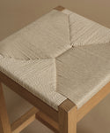 Close-up of a wooden stool with a woven rope seat in a light beige color, set against a solid warm beige background.