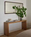 A minimalist Malibu Console Table in white oak with box joint details displays stacked books, a stone sculpture, and a tall vase of green branches. Above it, a large rectangular mirror hangs on the white wall.
