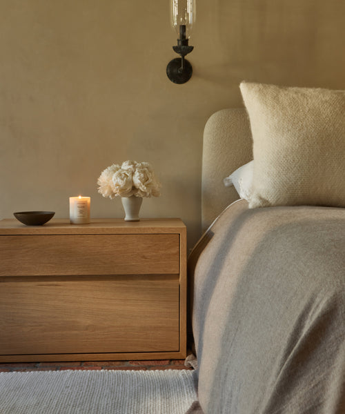 A cozy bedroom features a light wood nightstand with a Mandeville Glass Candle, a bowl, and white flowers next to a bed with beige bedding, all set against a neutral wall and illuminated by a wall sconce above.