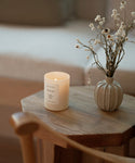 The Mandeville Glass Candle, hand-poured in Los Angeles, and a beige vase with dried flowers rest on a wooden table, with a blurred sofa behind them for a cozy, minimalist look.