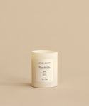 A white Mandeville Glass Candle in a minimalist container rests on a beige surface. Its label lists Lemon, Eucalyptus, and Vervain scents and notes that its hand-poured in Los Angeles.