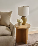 A Marin Side Table stands next to a beige sofa, displaying a ceramic lamp with a white shade and a vase of white flowers. The space is decorated in neutral tones with calm, minimalist style.