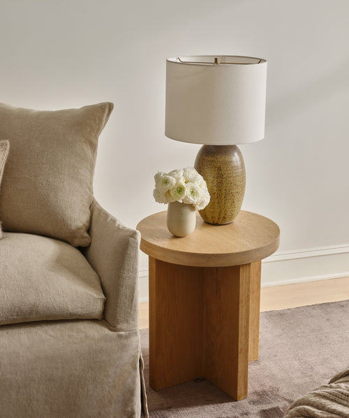 A Marin Side Table stands next to a beige sofa, displaying a ceramic lamp with a white shade and a vase of white flowers. The space is decorated in neutral tones with calm, minimalist style.