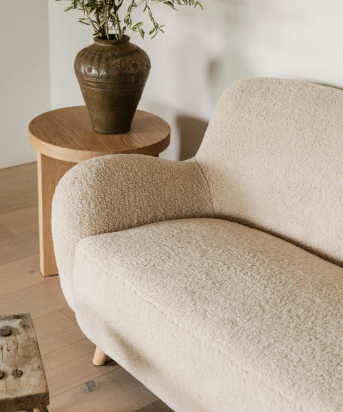 A cozy, textured beige sofa sits next to the minimalist Marin Side Table, topped with a brown ceramic vase of green leafy branches. The light, minimal setting is completed by a rustic wooden coffee table in front.