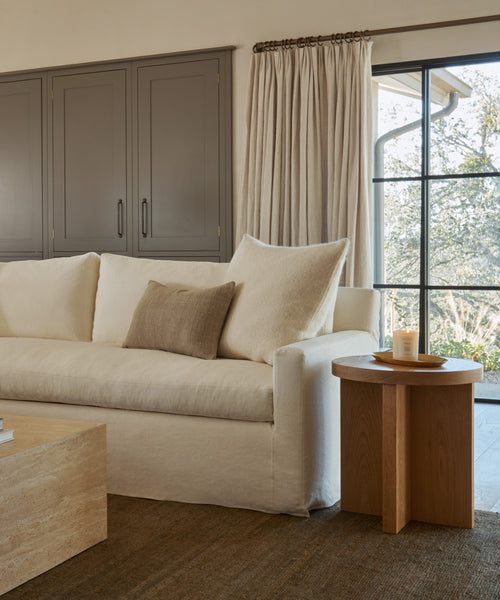 A cozy living room with a white sofa, beige pillow, the Marin Side Table holding a candle, a light wood coffee table, brown cabinets, beige curtains, and large windows revealing trees outside.