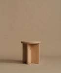 A minimalist light natural wood side table with three intersecting vertical legs, set against a solid warm beige background.