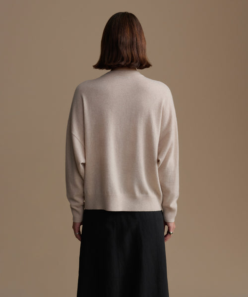 Woman wearing a beige cashmere sweater and dark pants against a brown background