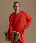 Woman wearing a red cashmere sweater and pants holding a beige bag against a plain background