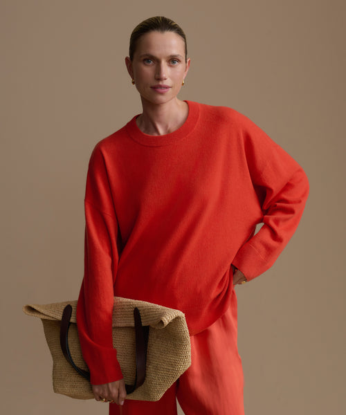 Woman wearing a red cashmere sweater and pants holding a beige bag against a plain background