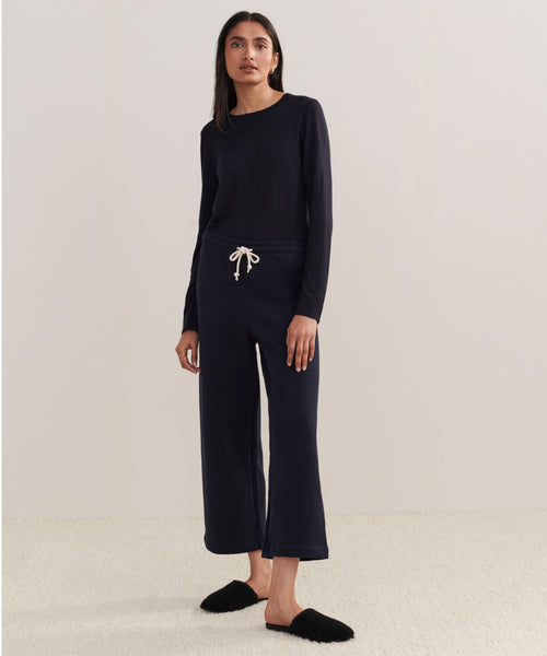 A woman stands against a plain background wearing the Marina Sweatpant—a matching dark navy, 100% cotton wide-leg sweatpant with a drawstring waist and long-sleeve top—paired with black slippers.