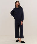 A woman stands indoors in cozy loungewear: navy blue 100% cotton Marina Sweatpant and matching sweatshirt, paired with black fuzzy slippers, against a plain light-colored background.