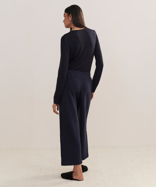 Someone stands facing away, wearing a dark long-sleeve top and the Marina Sweatpant—100% cotton, wide-leg loungewear pants—with black slippers, on a light carpet against a plain background.