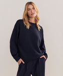 A woman with blonde hair wears a loose-fitting, dark navy Marina Sweatshirt and matching pants, styled as boxy loungewear, standing with hands in her pockets against a plain light backdrop.