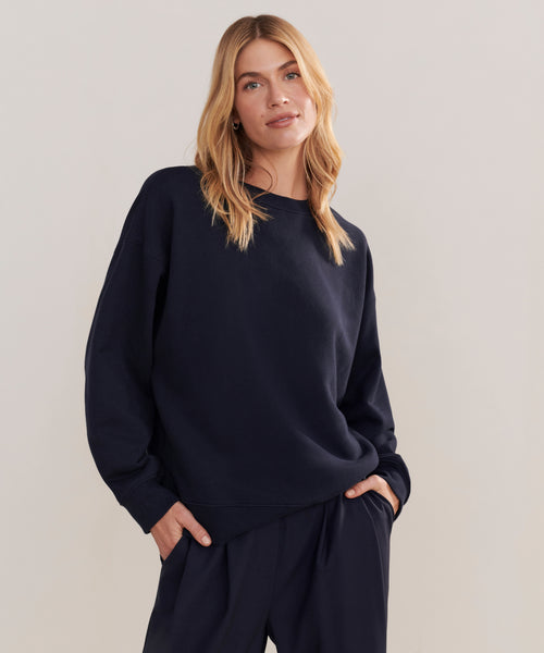 A woman with blonde hair wears a loose-fitting, dark navy Marina Sweatshirt and matching pants, styled as boxy loungewear, standing with hands in her pockets against a plain light backdrop.
