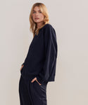 A woman with long blonde hair models the loose-fitting, dark navy Marina Sweatshirt and matching pants, standing with her hands in her pockets against a plain light background.