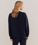 A person with long, blonde hair is seen from behind wearing a loose-fitting Marina Sweatshirt in dark navy, paired with matching loungewear pants, standing against a plain light background.