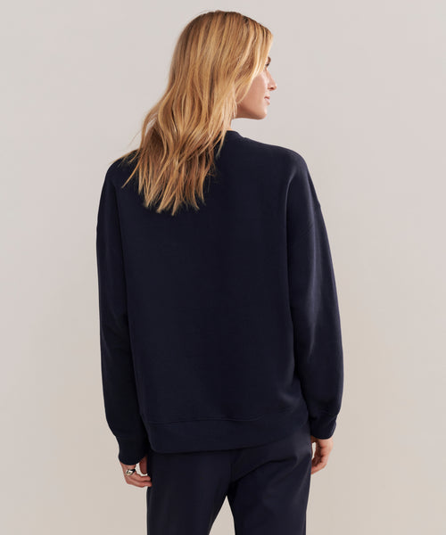 A person with long, blonde hair is seen from behind wearing a loose-fitting Marina Sweatshirt in dark navy, paired with matching loungewear pants, standing against a plain light background.