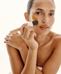 A woman with bare shoulders uses the Mask Brush to apply a restorative mask to her cheek, gazing calmly at the camera. Her hair is pulled back, creating a serene, natural look that captures an at-home spa experience.