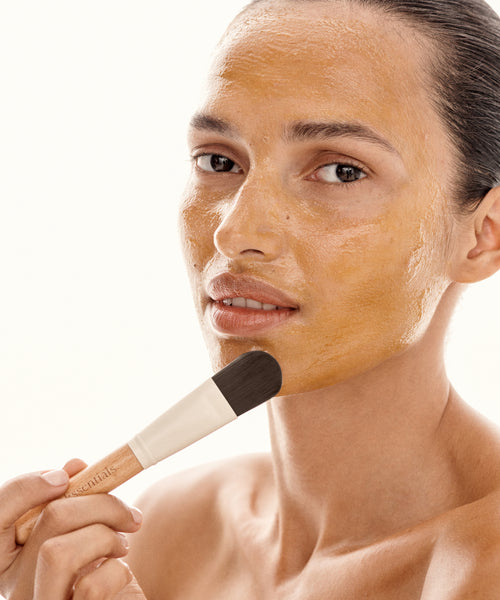 With hair pulled back, a person uses the Mask Brush to apply a golden-yellow restorative mask to smooth skin, looking neutrally at the camera and creating an at-home spa vibe against a plain, light background.