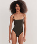 A woman with voluminous curly hair wears the Petite Square Maillot Swimsuit, a dark square-neck, spaghetti-strap style made from recycled nylon swimwear, standing against a plain light background.