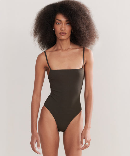 A woman with voluminous curly hair wears the Petite Square Maillot Swimsuit, a dark square-neck, spaghetti-strap style made from recycled nylon swimwear, standing against a plain light background.