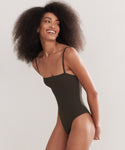 A woman with voluminous curly hair smiles and looks to the side while wearing a fitted dark green Petite Square Maillot Swimsuit, made from recycled nylon, set against a plain light background.