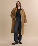 Against a plain beige background, a person wears the McCall Trench Coat—long, tan, water resistant, crafted from Italian fabric—paired with a black t-shirt, dark gray jeans, and black boots. One hand rests in their coat pocket; their expression is relaxed.