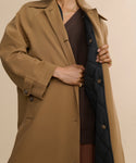 Someone models the McCall Trench Coat, a water-resistant tan coat with a dark quilted lining, holding it open to reveal a brown sweater underneath. Gold necklace and rings complete the look; their face remains unseen.