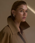 A woman with slicked-back hair and hoop earrings models the tan, water-resistant McCall Trench Coat with a high collar, looking thoughtfully to the side against a neutral background with a beam of light.