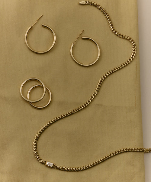 Two pairs of 14k gold Medium Eloise Hoops and a gold chain necklace with a rectangular clasp from Jenni Kayne jewelry are displayed on a light gold fabric background, creating an elegant, heirloom-worthy arrangement.