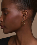 A close-up side profile of a dark-skinned woman with her hair pulled back, wearing the Medium Eloise Hoops in 14k gold and a black top, set against a neutral background that highlights the heirloom-quality jewelry.