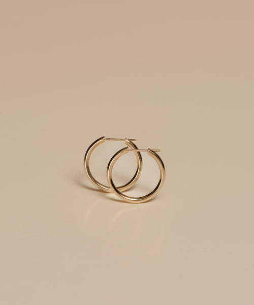 The Medium Eloise Hoops from Jenni Kayne Jewelry, crafted in simple 14k gold, are displayed upright against a smooth, light beige background.