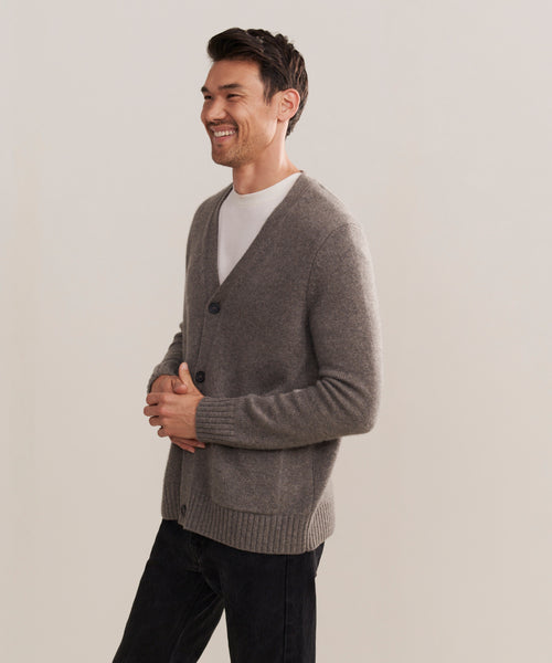 A man smiles against a plain background, wearing a Mens Cashmere Cardigan in grey over a white t-shirt and black pants, his hands loosely clasped in front of him.