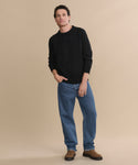A man stands against a beige background wearing the Men's Cashmere Fisherman Sweater in black, paired with blue jeans and brown shoes. He has one hand in his pocket and a relaxed expression.