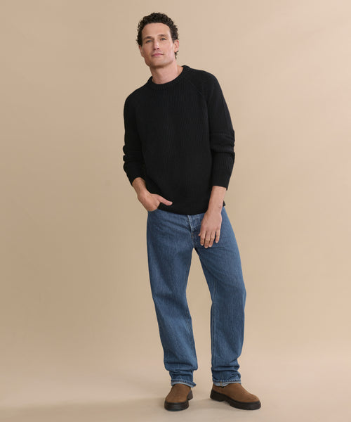 A man stands against a beige background wearing the Men's Cashmere Fisherman Sweater in black, paired with blue jeans and brown shoes. He has one hand in his pocket and a relaxed expression.