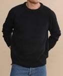 A person models the Men's Cashmere Fisherman Sweater in black with blue jeans, standing against a beige background, hands behind their back and head partially out of frame.