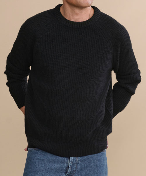 A person models the Men's Cashmere Fisherman Sweater in black with blue jeans, standing against a beige background, hands behind their back and head partially out of frame.