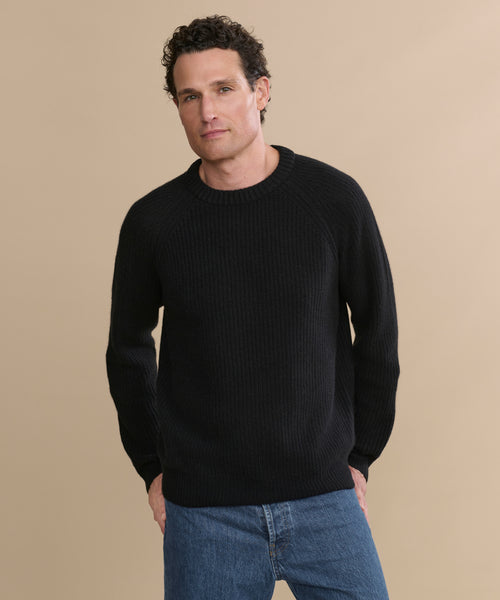 A man with short curly hair wears the Men's Cashmere Fisherman Sweater in black and blue jeans, standing against a plain beige background and looking at the camera with a relaxed expression.