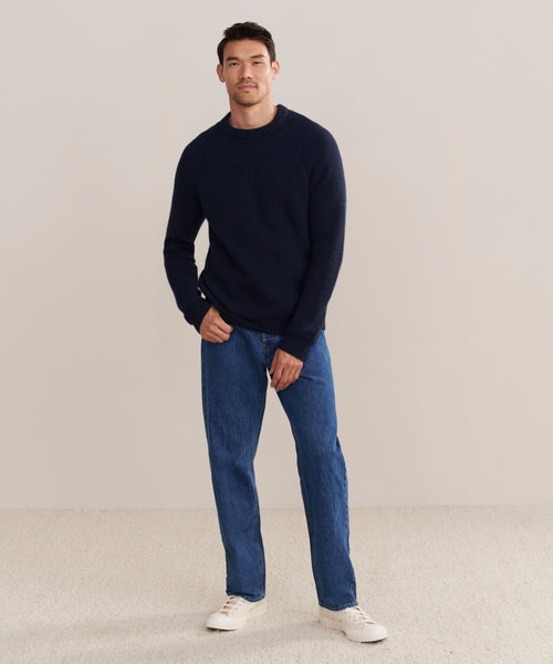 A man with short dark hair and a slight smile stands against a beige background, wearing a navy Mens Cashmere Fisherman sweater, blue jeans, and white sneakers. One hand is in his pocket.