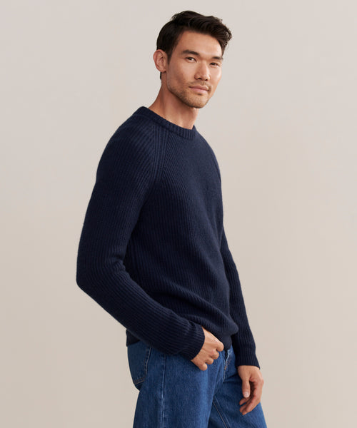 A man with short dark hair wears the Mens Cashmere Fisherman sweater in navy and blue jeans, standing against a plain light background and looking slightly to the side with a relaxed expression.